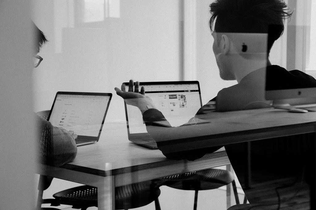 Zwei Personen in einem modernen Büro besprechen Inhalte auf ihren Laptops; einer zeigt mit der Hand auf den Bildschirm, der andere hört zu. Die Szene ist durch eine Glasscheibe fotografiert und wirkt durch Reflexionen leicht doppelt.