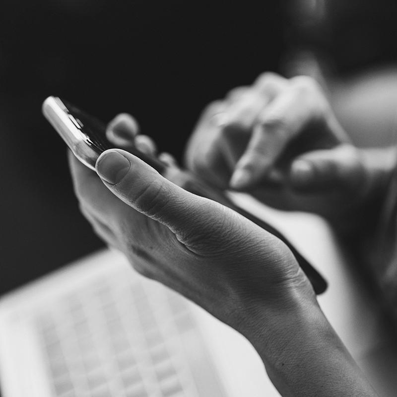 Schwarz-weiß-Aufnahme von zwei Händen, die ein Smartphone bedienen, im Hintergrund ist unscharf eine Tastatur erkennbar